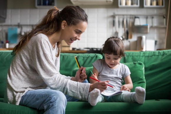 Smiling nanny and preschool kid girl drawing with colored pencils sitting on sofa together