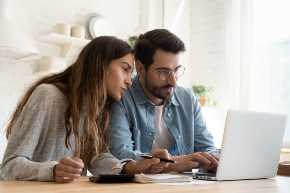 Couple financial planning while sitting at kitchen table looking at something on a laptop and taking notes.