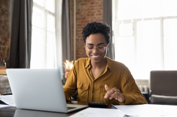 Woman sitting with her laptop and looking happy at a calculator.