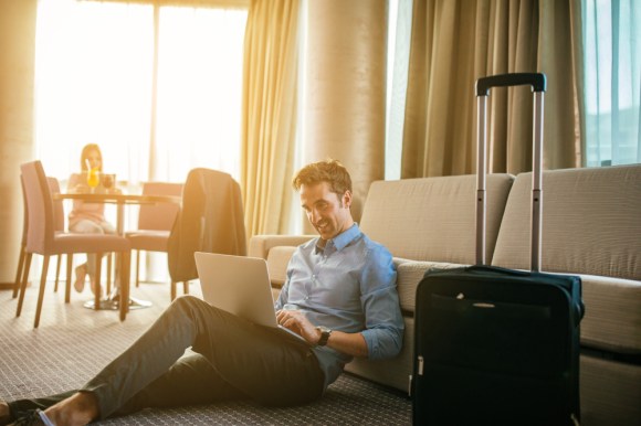 Man working in a hotel room with a woman sitting in the background.