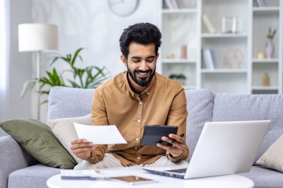 Young man sitting on his couch smiling while handling documents.