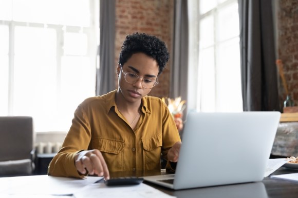 Young Black woman using a calculator and typing on her laptop.